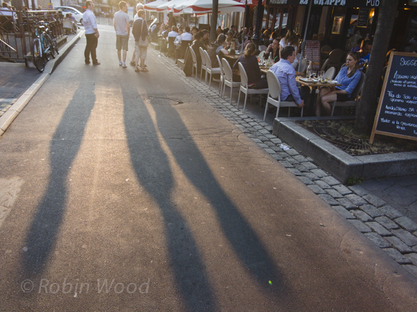 Pedestrians cast long shadows walking by packed cafes.